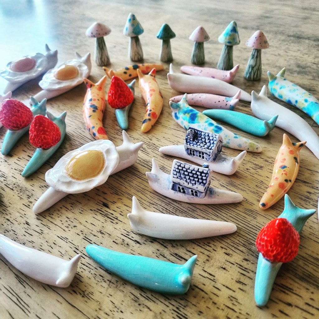 A variety of colorful ceramic snail figurines are arranged on a wooden surface, including some with fruit and small houses on their backs.