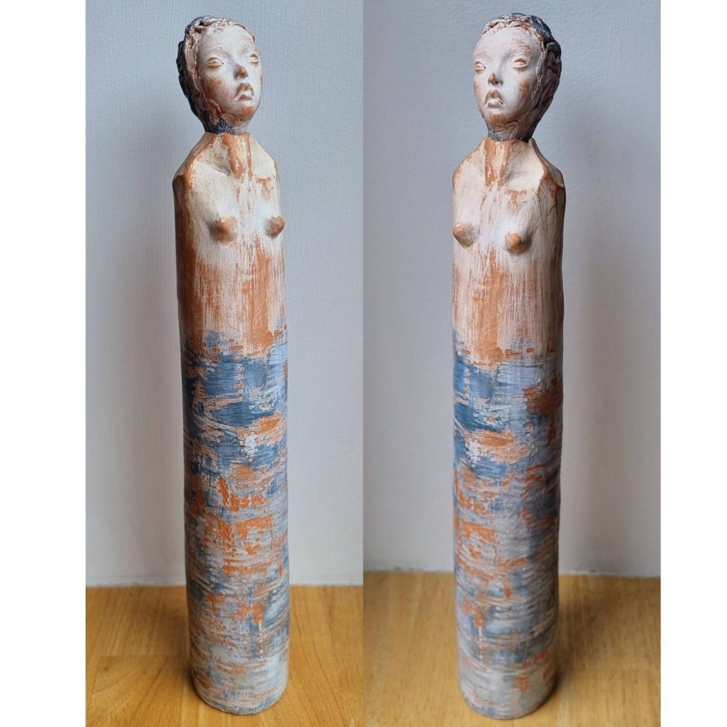 A tall, slender ceramic sculpture of a figure with a stylized face and body. The sculpture features a textured surface with a blend of earthy colors, including orange and blue, showcasing the beauty of accidental mark making in ceramics.